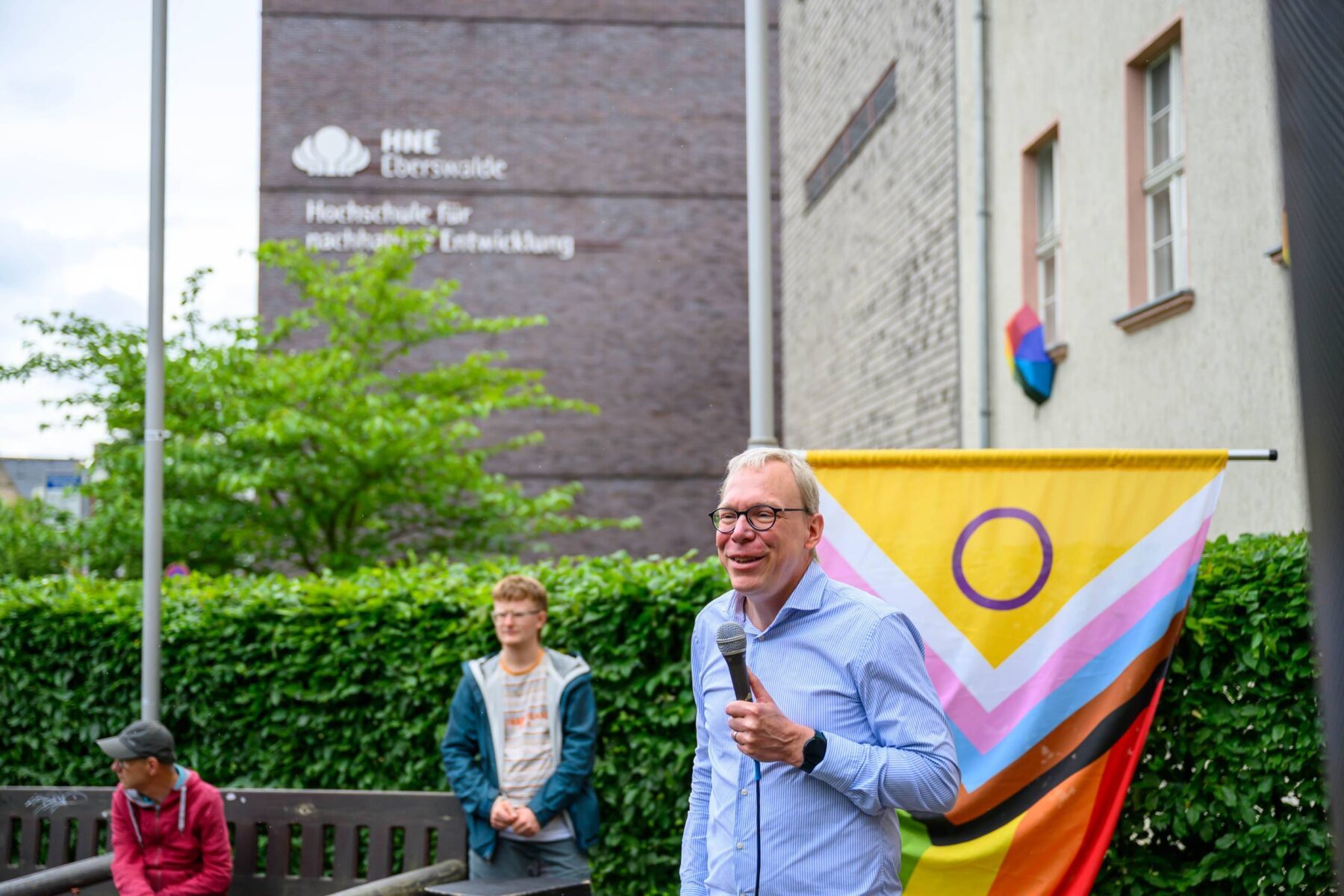 Markus Koenecke, Kanzler der Hochschule für nachhaltige Entwicklung Eberswalde (HNEE) eröffnet mit einem Mikro in der Hand die Queeren Wochen 2025 in Eberswalde, Progress-Pride-Flagge im Hintergrund.