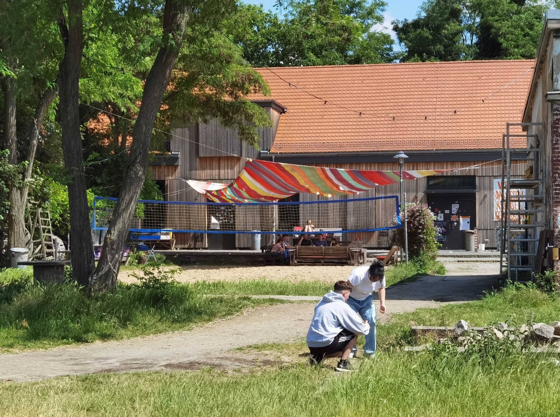 Zwei Personen vor einem Gebäude mit bunter Markise und Volleyballnetz, umgeben von grüner Natur und sonnigem Wetter.